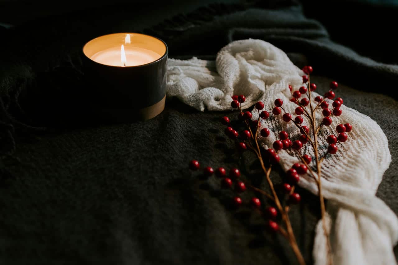 A lit candle in a dark ceramic holder beside a cream knit throw and sprigs of red winter berries.