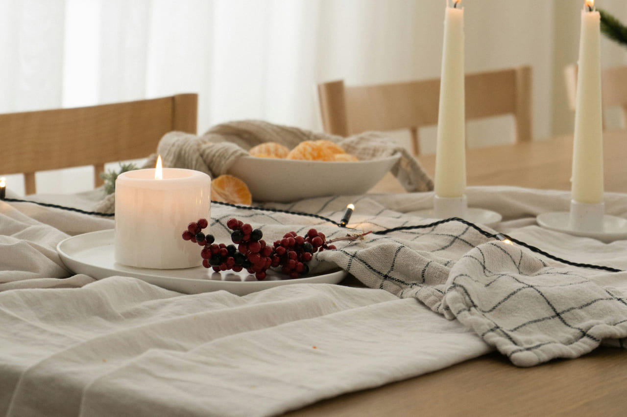 A linen-covered table by a sunlit window with taper candles, a pillar candle and winter berries.