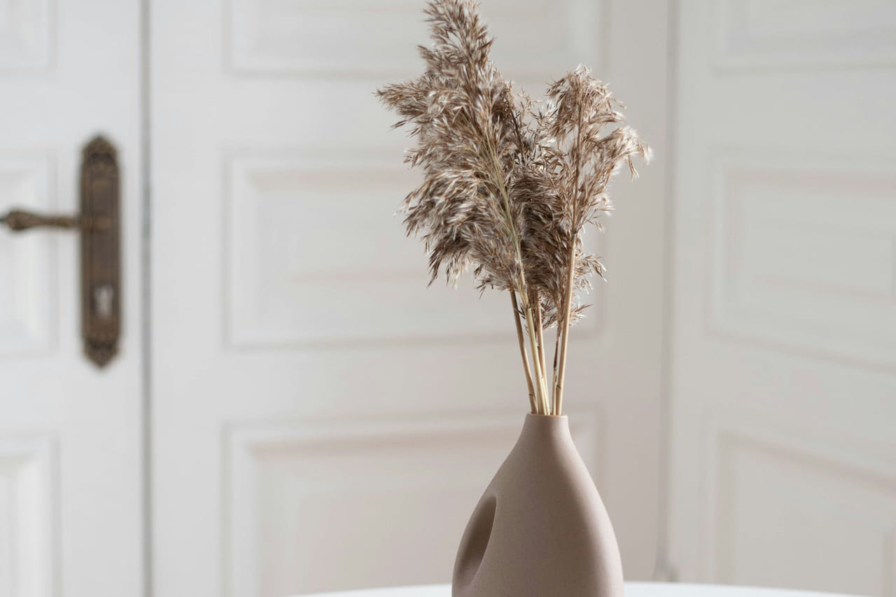 A matte clay vase of dried grasses on a round white table before a paneled door.