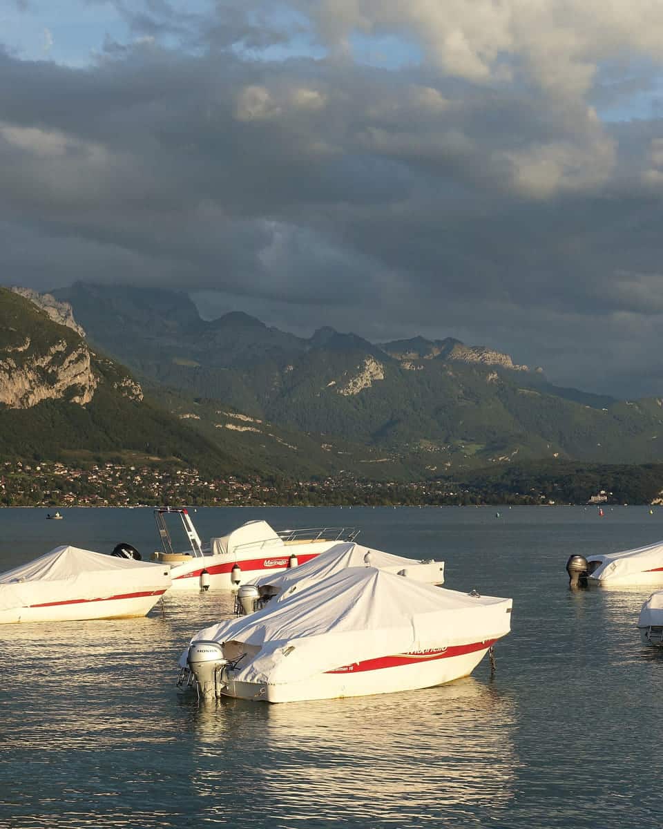 White boats moored on a calm, grey-green lake beneath dark, cloud-topped mountains.