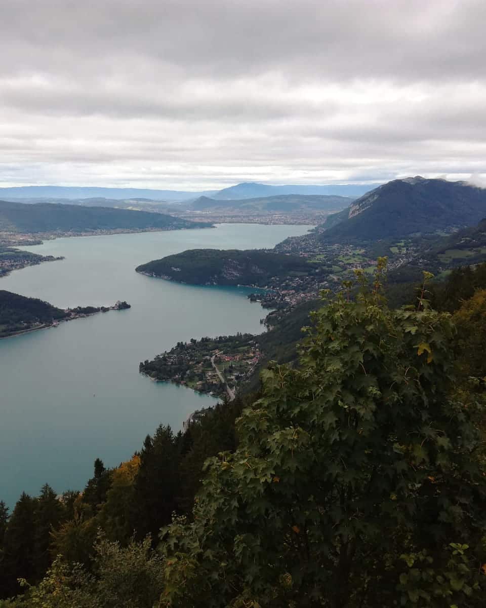 Lake Annecy seen from high above at the Col de la Forclaz, the water winding between forested mountains under a pale sky.
