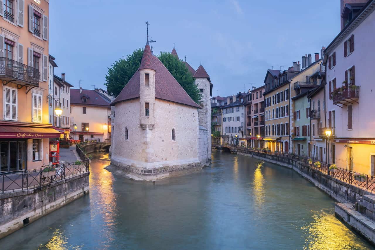The Palais de l’Isle, a stone fortress on an islet in the Thiou canal, at dusk, with lit quays and their reflections.