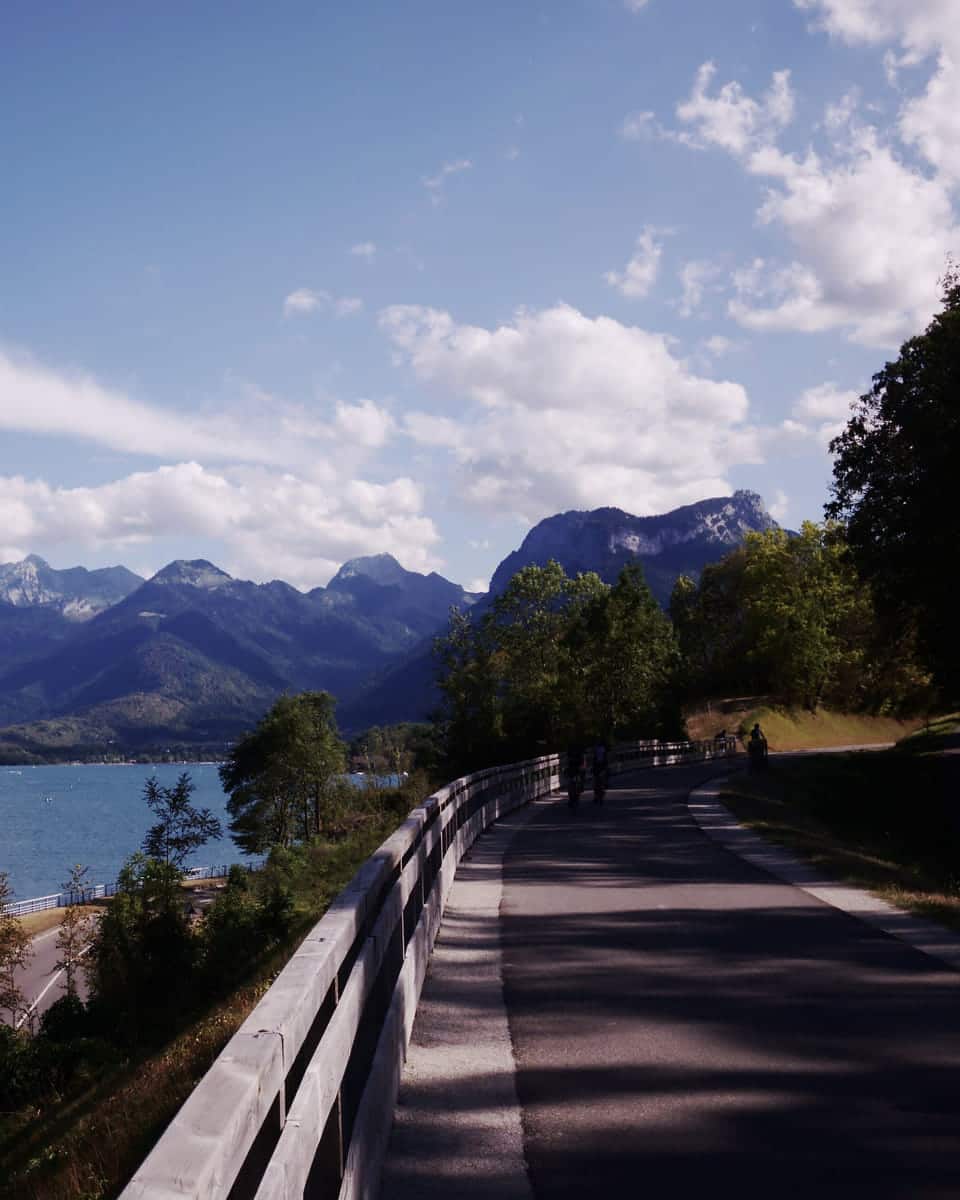 A paved cycle path curving along the lakeshore toward a ring of mountains, the water deep blue on the left.