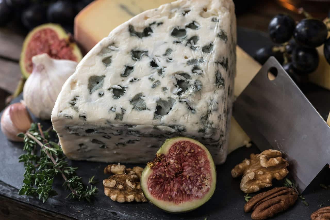 A wedge of blue-veined cheese with fresh figs, walnuts, garlic and thyme on a dark board.