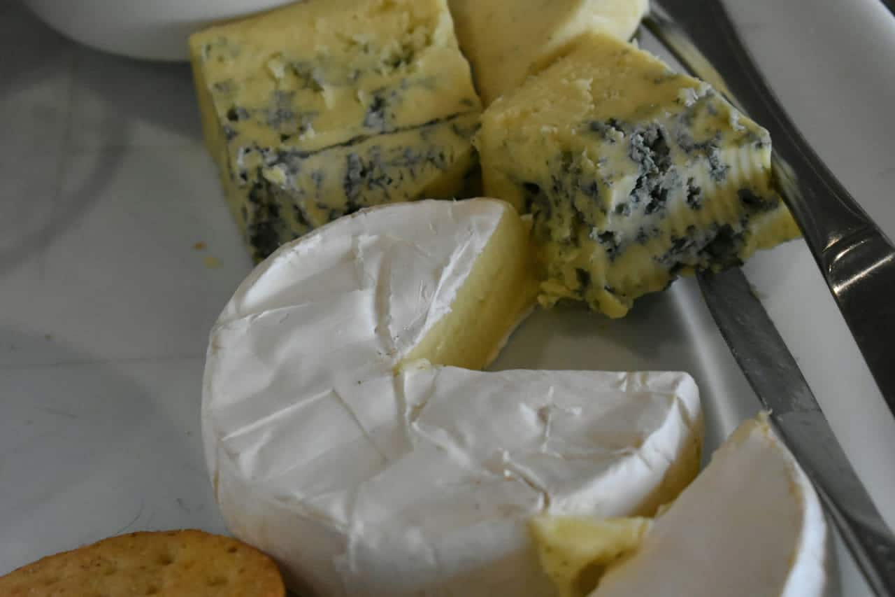 A round of soft white-rind cheese, cut to show its creamy centre, beside crumbly wedges of blue cheese and a cheese knife.