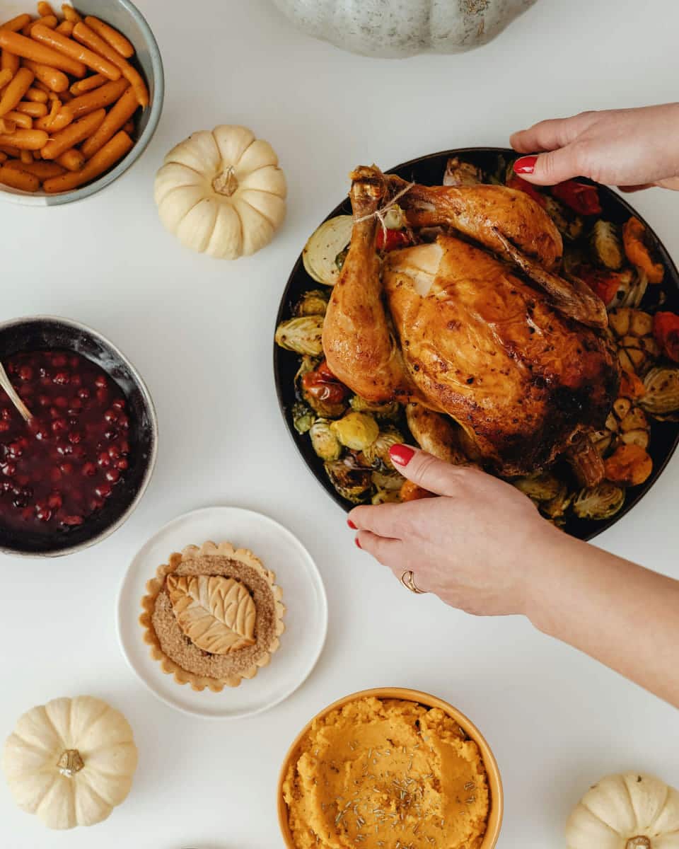 Two hands setting a platter of roast turkey and vegetables on a white table among bowls of cranberry sauce, mashed sweet potato and small white pumpkins.