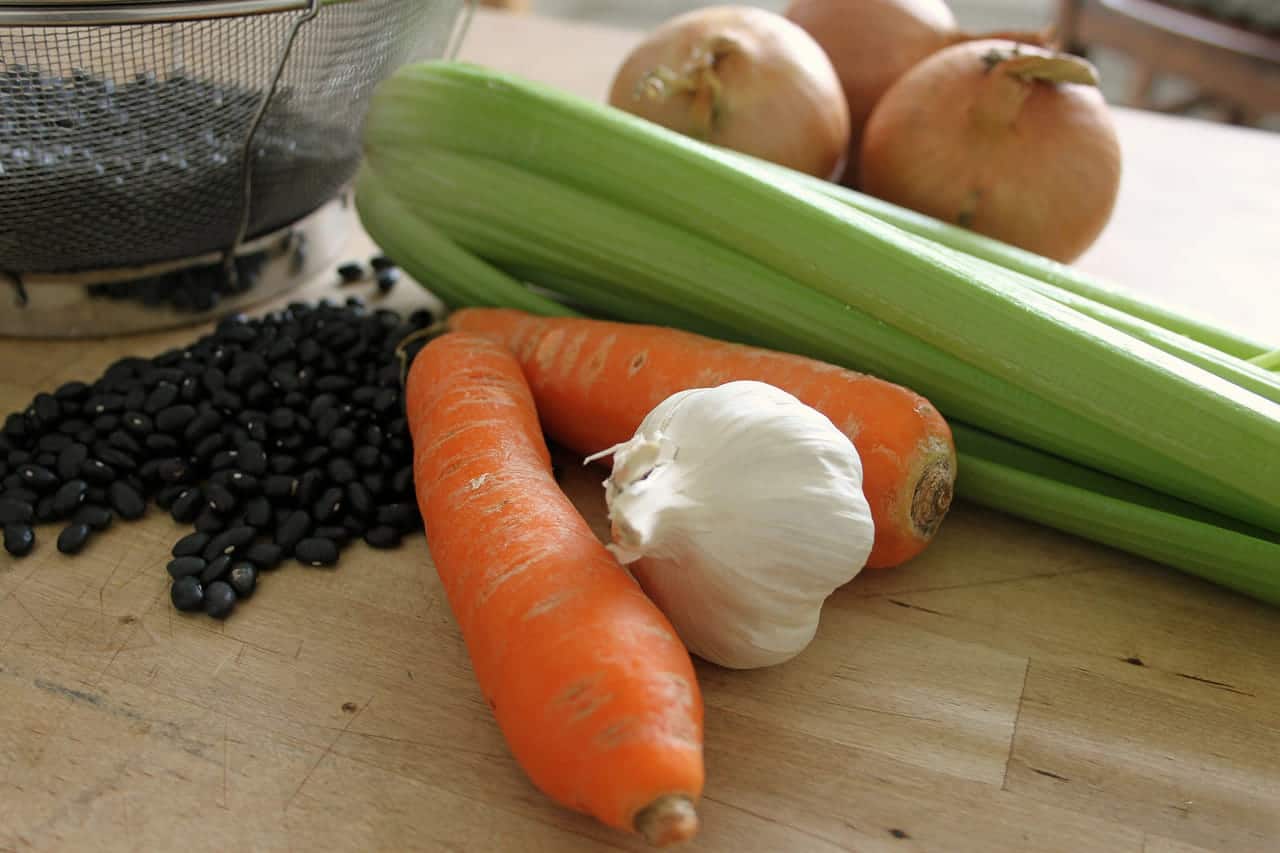 Whole carrots, a head of garlic, celery stalks and yellow onions on a wooden chopping board, beside a scatter of dried black beans.
