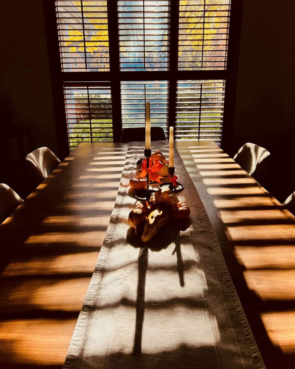 A long wooden dining table in late sun through tall shuttered windows, dressed with a pale linen runner, two white tapers and a low cluster of small squash and gourds.