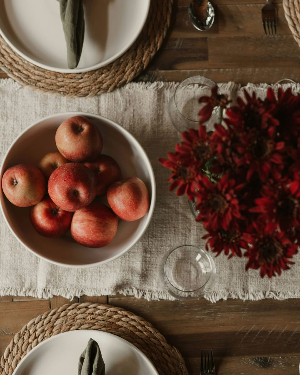 Overhead view of a dressed dinner table with a linen runner, apples and dark flowers.